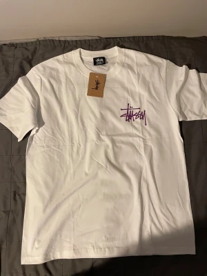 Vit Stüssy t-shirt med lila logga XL - Vit t-shirt från Stüssy i storlek XL med ikonisk lila Stüssy-logga tryckt på bröstet och stort på ryggen. Klassisk passform och rund hals. Tillverkad i mjuk bomull, perfekt för en clean streetwear-look.