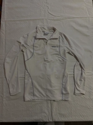 Vit långärmad träningströja Craft - Säljer en vit långärmad träningströja från Craft i storlek S. Tröjan har half zip framtill, broderad logga på bröstet och kontrastsömmar i grått. Perfekt för sport och utomhusaktiviteter, tillverkad i mjukt syntetmaterial som andas.