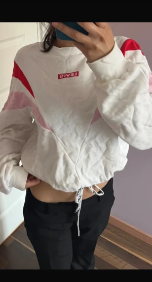 Vit och röd Levi's sweatshirt XS - Säljer en vit sweatshirt från Levi's i storlek XS med röda och rosa detaljer på axlarna och bröstet. Tröjan har lång ärm, snörning med logotyp vid midjan och en croppad passform. Materialet är mjuk bomull och polyester, perfekt för en chill look.