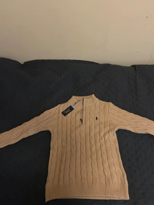 Beige stickad half zip från Polo Ralph Lauren - Snygg beige tröja från Polo Ralph Lauren med half zip och kabelstickat mönster. Tröjan har ribbad krage och muddar, samt det klassiska Polo-logot broderat på bröstet. Perfekt för dig som gillar stilrena och tidlösa plagg. Tröjan har storleken L men känns som M därav säljer jag den !!!