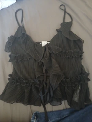 Svart volang linnetopp från Amisu XXS - Säljer en svart linnetopp från Amisu i storlek XXS. Toppen har tunna axelband, plisserat tyg och flera lager med volanger. Den har snörning framtill och är croppad i modellen, perfekt för dig som gillar detaljer och en feminin look.