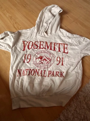 Vit hoodie Yosemite från Kappahl - Vit hoodie från Kappahl med rött tryck 'Yosemite National Park 1991' på framsidan. Tröjan har huva, ribbad nederkant och är tillverkad i mjuk bomull. Perfekt för dig som gillar coola tryck och avslappnad stil.