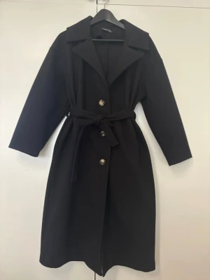 Svart kappa med bälte från Boohoo - Stilren svart kappa från boohoo med klassisk krage, knappar framtill och ett matchande bälte i midjan. Kappan är i strl 36 och på sista bilderna kan ni se att de finns små hål i fickorna utöver detta jätte fin kappa
