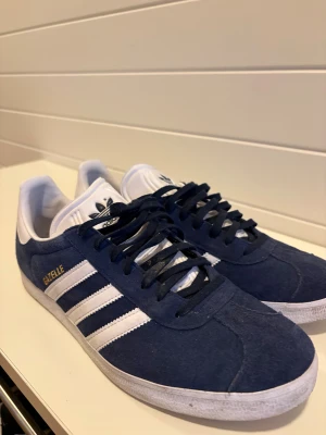 Adidas Gazelle blå sneakers strl 44 - Säljer ett par Adidas Gazelle sneakers i marinblå mocka med vita detaljer och klassiska tre ränder på sidan. Skorna har snörning, platt sula och rund tå. Använda cirka 10 gånger max och har bara stått här och inte används. Perfekt för dig som gillar retrovibe och snygg streetstil.