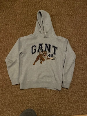 Grå hoodie med broderi från GANT - Snygg grå hoodie från GANT med stort broderi på bröstet, där en tiger och texten 'GANT 49 EST.' syns. Tröjan har huva, långa ärmar och ribbade muddar. Perfekt för dig som gillar sportig och cool stil.