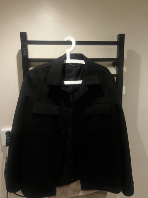 Svart Overshirt  - Säljer nu min svarta trendiga overshirt med klassisk krage och stora bröstfickor. Jackan har knäppning framtill och raka ärmar. Den är  från Zara i storlek S. Den har en tunn men ändå varm ock tjock känsla vilket gör den perfekt för båda inomhus och utomhusanvändning! Först till kvarn gäller!