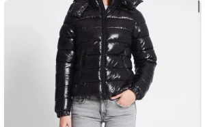 Svart glansig pufferjacka med huva - Säljer en svart, glansig pufferjacka med huva och dragkedja framtill. Jackan har quiltad design, två sidofickor med dragkedja och långa ärmar. Perfekt för kalla dagar när du vill hålla stilen och värmen.