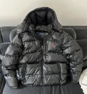 Svart pufferjacka från Polo Ralph Lauren - Snygg svart pufferjacka från Polo Ralph Lauren med glansig finish och klassisk röd logga på bröstet. Jackan har huva, flera dragkedjeförsedda fickor och en rejäl, vadderad känsla som håller dig varm under kalla dagar.