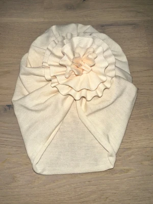 Beige turban med volangdetalj Beige mössa - Snygg beige turban med stor volang framtill som ger en cool och unik look. Mjuk och stretchig, perfekt för dig som vill sticka ut lite extra. Materialet känns som bomull och passar skönt på huvudet. Perfekt accessoar för att lyfta din outfit.En söt turbanmössa med en stor blomma på toppen. Den har en ljus, neutral färg. För barn 1 år +/-  stretch material super bra!👍🏻 