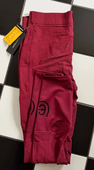 Nya Vinröda ridbyxor Equestrian Stockholm med tags kvar - Snygga vinröda ridbyxor från Equestrian Stockholm med silikongrepp och praktisk ficka på benet. Byxorna har hög midja och är tillverkade i ett stretchigt material som ger bra rörelsefrihet. Perfekta för dig som vill ha stil och funktion i stallet. Helt nya med lapparna kvar. 