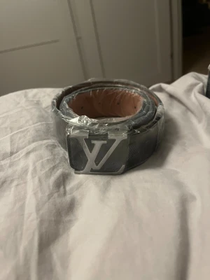 Svart rutigt bälte - Säljer ett stilrent svart bälte, rutigt mönster och en stor silverfärgad logga som spänne. Bältet är tillverkat i skinn med en snygg finish och har en brun insida. Perfekt för dig som vill ha en lyxig accessoar med statement.