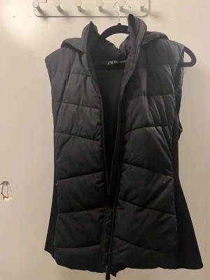 Svart quiltad dunväst från Zara - Svart dunväst från Zara med quiltad design och huva. Västen har dragkedja framtill och är figursydd. Strlk 36
