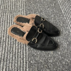 Svarta loafers med päls från Zara - Säljer ett par svarta loafers från Zara med beige fuskpäls på insidan och snygga metallspännen framtill. Skorna har spetsig tå och platt sula, vilket ger en cool och trendig look. Perfekt för dig som gillar att sticka ut med dina skor.
