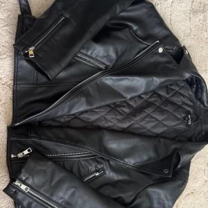 Svart skinnjacka från Fórmula Joven XL - Snygg svart skinnjacka från Fórmula Joven i bikerstil med quiltat foder. Jackan har flera silvriga dragkedjor, klassisk krage med tryckknappar och detaljerade sömmar. Perfekt för dig som vill ha en edgy look med stilrena detaljer. Den må va XL men den sitter som en M då den e ganska liten i storleken. 
