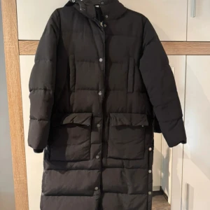 Svart lång pufferjacka med huva - Lång svart pufferjacka med huva och stora fickor framtill. Jackan har tryckknappar längs hela framsidan och på sidorna, samt quiltad design för extra värme. Perfekt för kalla dagar och har en stilren look.