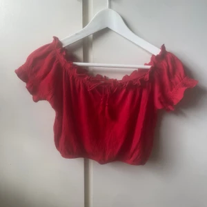 Röd croppad blus med volang - Säljer en röd croppad blus med offshoulder och korta ärmar. Toppen har volangdetaljer runt halsringningen och ärmarna samt en snygg knytning framtill. Lappen sitter tyvärr inte kvar så vet inte exakt storlek och vart den är ifrån☺️