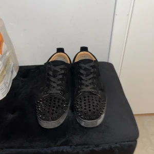 Svarta Loubs med nitar - Säljer ett par svarta sneakers från Christian Louboutin med coola svarta nitar på tån och klassisk röd sula. Skorna har snörning, rund tå och är tillverkade i mocka med läderfoder. Perfekt för dig som vill sticka ut med en edgy vibe.