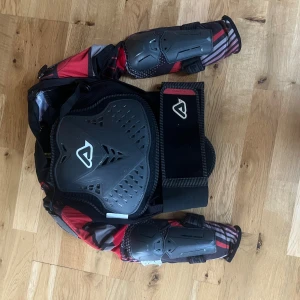 Acerbis Scudo Junior 2.0 skyddsväst L/XL - Skyddsväst från Acerbis, modell Scudo Junior 2.0, i storlek L/XL. Västen har robusta svarta och grå skyddsplattor på bröst och rygg, samt armbågsskydd. Designen är sportig med röda och grå detaljer och randigt mönster på ärmarna. Perfekt för motocross eller liknande aktiviteter.