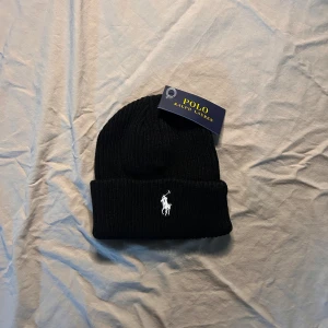 Svart ribbad mössa från Polo Ralph Lauren - Svart ribbstickad mössa från Polo Ralph Lauren med klassisk broderad logga framtill i vitt. Tjockt uppvik och stilren design som passar perfekt till kyliga dagar. Materialet är mjukt och värmande, och mössan har en enkel, tidlös look. Pris kan diskuteras vid snabb affär och varan skickas samma dag!