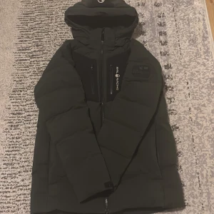 Svart dunjacka från Sail Racing - Svart dunjacka från Sail Racing med huva och snygga svarta detaljer på bröstet och ryggen. Jackan har flera dragkedjor framtill och patch på ärmen. Fylld med Allied Feather + Down, 500 fill power. Perfekt för kalla dagar och har en clean, sportig look.Har lite slitage/hål men inget som inte kan laggas.