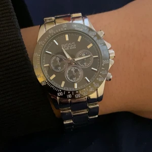 Silverfärgad kronografklocka från Hugo Boss - Stilren kronografklocka från Hugo Boss i silverfärgat rostfritt stål. Klockan har en rund urtavla med flera små visare, markerade index och tachymeter på ringen. Länken är robust och har ett snyggt Boss-graverat spänne. Perfekt för dig som gillar klassisk design med sportig touch.