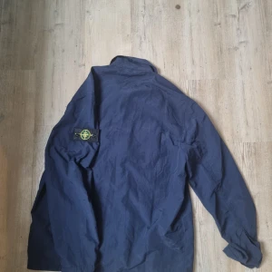 Blå overshirt från Stone Island - Säljer en mörkblå overshirt från Stone Island med klassisk patch på vänster ärm. Jackan har två bröstfickor med lock och hel dragkedja framtill. Materialet är lätt och syntetiskt, perfekt för lager-på-lager. Stilren och enkel design som funkar till många outfits. 