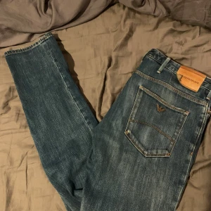 Emporio Armani blå jeans  - Snygga blå jeans från Emporio Armani med klassisk femficksdesign och logga på bakfickan. Jeansen är tillverkade i slitstarkt denim med coola slitningar och en liten reva på ena benet. Perfekt för dig som gillar stilrena och trendiga jeans. ena fickan är hål i andra är sydd därav priset