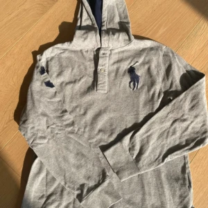 Grå hoodie från Polo Ralph Lauren - Snygg grå hoodie från Polo Ralph Lauren med stor marinblå logga på bröstet och knäppning vid halsen. Tröjan har huva med mörkblå insida och långa ärmar. Passar folk runt 160 😃