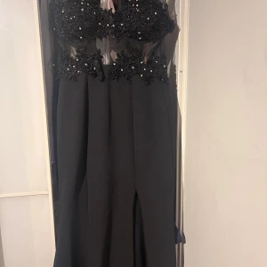 Svart långklänning med pärlbroderi - Svart långklänning med transparent mesh upptill och detaljerad pärlbroderad spets över byst och midja. Klänningen har v-ringad urringning och är ärmlös, med en elegant slits framtill. Perfekt för dig som vill sticka ut med glittriga detaljer och unik design.