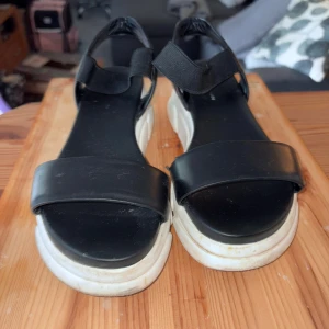 Svarta chunky sandaler med vit sula - Svarta sandaler med breda remmar och elastiskt band över foten. De har en chunky vit plattformssula som ger en cool och modern vibe. Materialet ser ut som syntet med en slät finish. Perfekt för dig som gillar streetstyle och vill ha något bekvämt och trendigt.