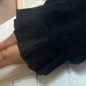 Svart plisserad kortkjol - Snygg svart plisserad kortkjol med klassisk midja och tydliga veck. Kjolen har en stilren look och passar perfekt till en stickad tröja eller crop top. Materialet känns mjukt och följsamt, och modellen ger en trendig siluett. Har inbyggda shorts