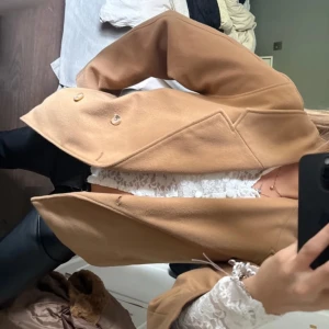 Beige kappa med knappar - Snygg beige kappa med dubbelknäppning och klassisk krage. Jackan har en rak passform och är tillverkad i ett mjukt material som ger en stilren look. Perfekt för dig som vill ha en tidlös och trendig jacka till höst och vinter.
