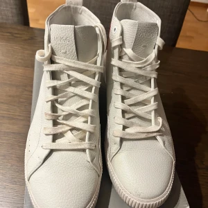 Vita high-top sneakers från Hugo - Snygga vita high-top sneakers från Hugo med röd logotyp på sidan och präglad Hugo-text på hälen. Skorna har snörning, rund tå och är tillverkade i strukturerat skinn med platt sula. Perfekt för dig som gillar stilrena och moderna sneakers med en exklusiv touch.