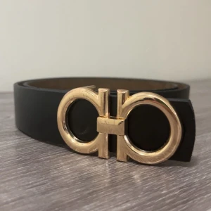 Svart skärp från Salvatore Ferragamo - Stilrent svart bälte i skinn från Salvatore Ferragamo med ikoniskt dubbel-Gancini-spänne i guld. Bältet har en klassisk, rak form och justerbara hål för perfekt passform. Perfekt accessoar för att lyfta din outfit med en lyxig touch.