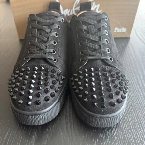 Christian Louboutin svarta sneakers med nitar - Svarta sneakers från Christian Louboutin i mocka med coola svarta nitar på tån och klassisk röd sula. Skorna har snörning och en rund tå, vilket ger en edgy och lyxig vibe. Perfekta för dig som vill sticka ut med exklusiva detaljer.