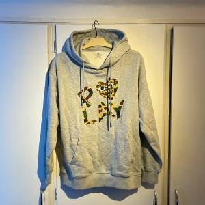 Grå hoodie PLAY med camo-tryck - Grå hoodie med huva och snörning, storlek M. Framsidan har ett stort PLAY-tryck i camouflagemönster med ett hjärta och ögon, inspirerat av streetwear. Mjuk och skön, perfekt för chill dagar. Priset är ej hugget i sten!