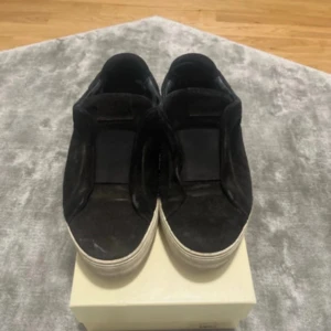 Svarta sneakers från Axel Arigato - Säljer ett par svarta sneakers från Axel Arigato med platt vit sula och rund tå. Skorna är tillverkade i mocka och har en stilren slip-on design utan snörning. Perfekta för dig som gillar minimalistisk och clean streetstil. Lite sne sula men annars bra skor, samt lite hål i hälen på insidan som de blir med alla skor, inte under sulan utan inuti men inget som märks eller speciellt syns!