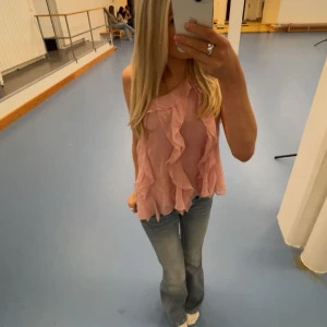 Rosa volang topp med oneshoulder - Säljer en superfin ljusrosa topp med volanger och oneshoulder-design. Toppen är ärmlös och har en luftig, flowy passform som känns riktigt somrig. Perfekt till jeans eller kjol för en söt och trendig look.