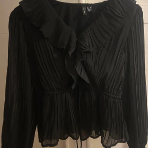  svart blus från Vero Moda - Säljer en svart blus från Vero Moda med volangkant längs ringningen och omlottkänsla framtill. Blusen har långa ärmar och resår i midjan för en snygg siluett. Tunn och luftig med feminina detaljer, perfekt för att lyfta din outfit.