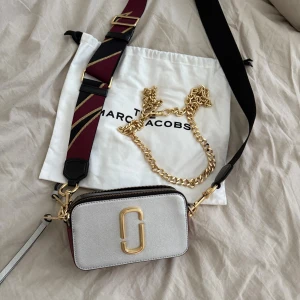 Marc Jacobs Snapshot axelväska silver - Snygg axelväska från Marc Jacobs i silverfärgat skinn med guldfärgade detaljer och logga framtill. Väskan har två fack med dragkedja, en bred randig axelrem i vinrött och svart samt en extra guldfärgad kedja. Perfekt storlek för det viktigaste.