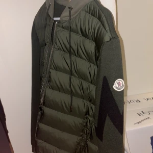 Grön Moncler pufferjacka med huva - Snygg mörkgrön pufferjacka från Moncler med stickade ärmar och huva. Jackan har dragkedja framtill, snörning vid huvan och Moncler-logga på ena ärmen. Ärmarna har ett svart zigzag-mönster som ger en cool kontrast. Perfekt för höst och vinter.