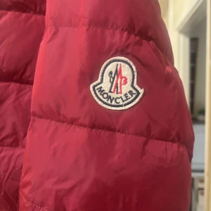 Röd Moncler dunjacka med huva - Säljer en röd dunjacka från Moncler med huva och grå stickad kant vid dragkedjan. Jackan har quiltad design, sidofickor med dragkedja och Moncler-logga på ärmen. Perfekt för kalla dagar och har en snygg, sportig look.