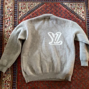Grå stickad tröja Louis Vuitton - Grå stickad tröja från Louis Vuitton med stort vitt LV-logo på bröstet. Klassisk rund halsringning och ribbade muddar vid ärmslut och nederkant. Perfekt för dig som vill ha en lyxig och stilren look.