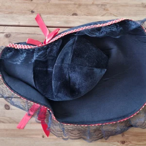 Svart pirathatt med rosetter - Svart pirathatt från Rubie's Costume Co med brett brätte dekorerat med svart spets och röd-guld broderad kant. Snygga röda rosetter på sidorna ger extra attityd. Perfekt för temafest eller maskerad! Den har legat oanvänd länge och kan behöva stärkas för att få formen att hålla sig. Kanske något för halloween?