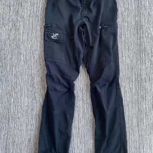 Svarta Nordwand cargopants RevolutionRace - Svarta Nordwand Pants från RevolutionRace med flera praktiska fickor, inklusive benficka med logga och dragkedja. Byxorna har normal passform och är tillverkade av slitstark polyester och bomull, perfekta för äventyr och outdoor. Snygg svensk design. Mått Midjan: ca 38 cm Längd från grenen och ner: ca 79 cm  96% nylon och 4% elastan  Kommer från från ett djur och rökfritt hem.