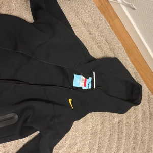 Svart NOCTA vindjacka Nike L - Svart vindjacka från Nike NOCTA i storlek L med huva och gul logga. Jackan har dragkedja, flera gula broderade detaljer och en ficka med vattentät dragkedja på ärmen. Snygg och sportig design som sticker ut med NOCTA-tryck. L passar som M