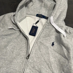 Grå zip från Polo Ralph Lauren - Snygg ljusgrå hoodie från Polo Ralph Lauren med dragkedja och klassisk broderad logga på bröstet. Tröjan har huva med vita snören, känguruficka och ribbade muddar. Perfekt för en chill och stilren look.