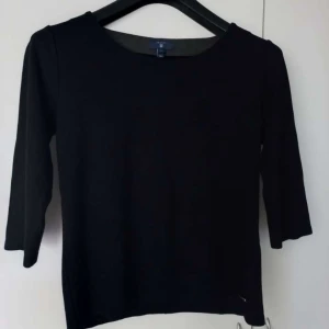 Svart topp med trekvartsärm från GANT - Svart stilren topp från GANT i storlek M med trekvartsärm och rund halsringning. Materialet är mjukt och stretchigt, troligen bomull med inslag av syntet. Diskret metallmärke med GANT-logga vid nederkanten ger en snygg detalj.