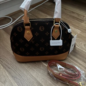 Mönstrad handväska från ZuobaoyouDai - Snygg handväska med klassiskt brunt och svart mönster, rundad form och guldfärgade detaljer. Väskan har två handtag och en avtagbar axelrem. Materialet är skinnimitation med bruna detaljer och dragkedja upptill. Perfekt för dig som gillar stilrena accessoarer.