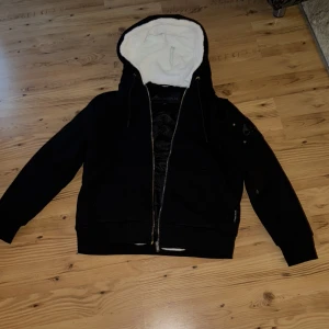 Moose knuckels bunny jacka - Svart hoodiejacka från Moose Knuckles med vit fluffig fuskpäls i huvan och quiltat svart innerfoder. Jackan har dragkedja framtill, snörning i huvan och en ikonisk logga på ärmen. Perfekt för kalla dagar och har en riktigt chill vibe.